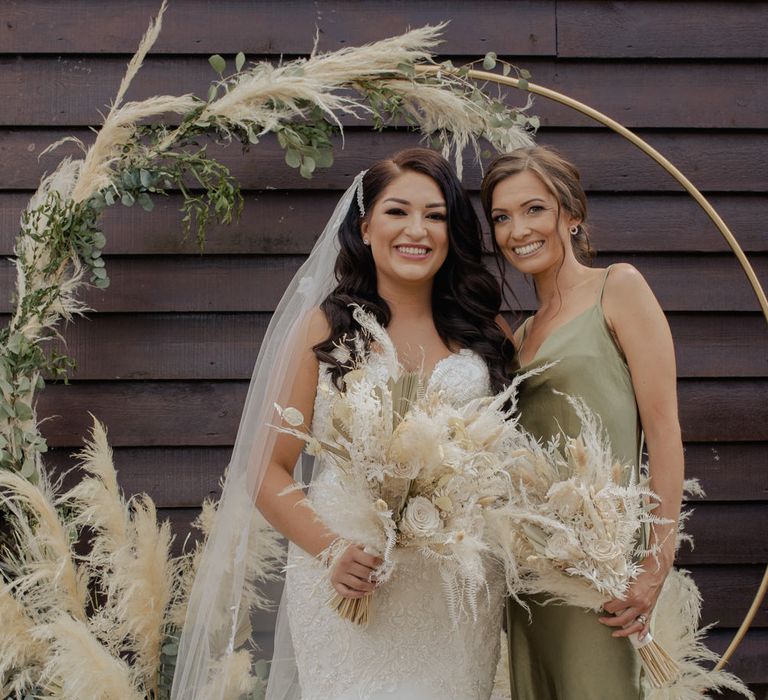 Bride in a fishtail wedding dress holding a dried flower wedding bouquet standing in front of a pampas grass hoop with her bridesmaid in a green satin dress