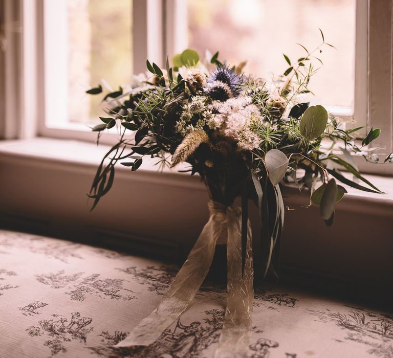 Blue and white wedding bouquet resting on a window sill with thistles and eucalyptus.