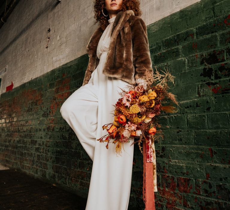 Bride in a wide leg bridal jumpsuit and brown faux fur coat holding an orange and yellow dried and fresh flower bouquet tied with ribbon