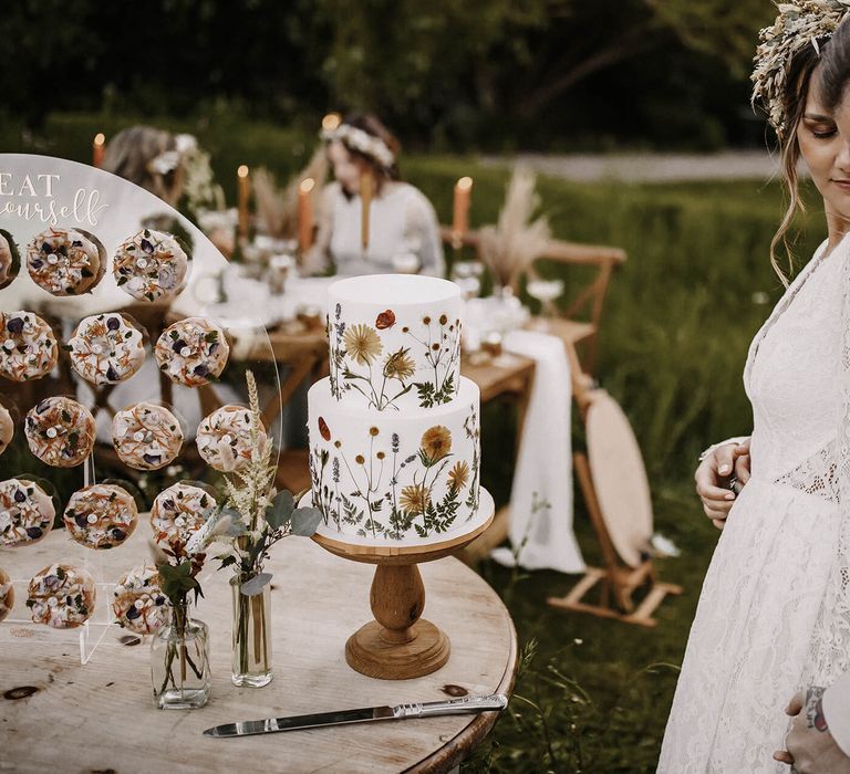 Groom in a beige suit embracing his bride in a lace wedding dress by their wedding cake table with doughnut wall and pressed flower cake 