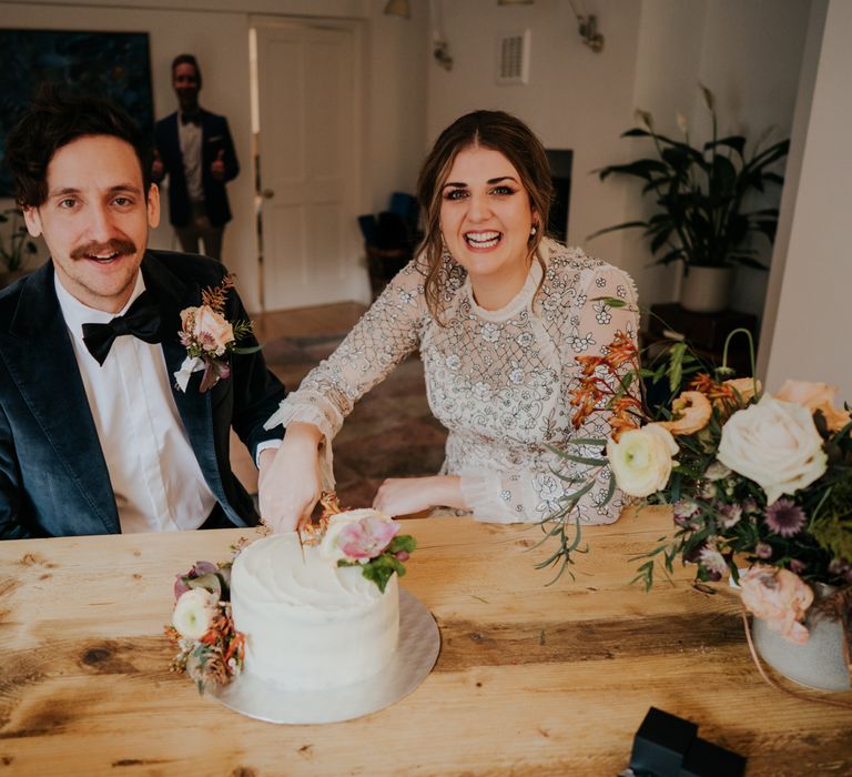 Bride & groom smile as they sit together and cut their wedding cake with floral decoration 