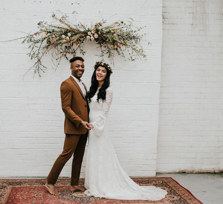 Bride and groom portrait in an industrial venue with groom in a brown suit, and bride in a lace dress standing on a rug under a flower installation