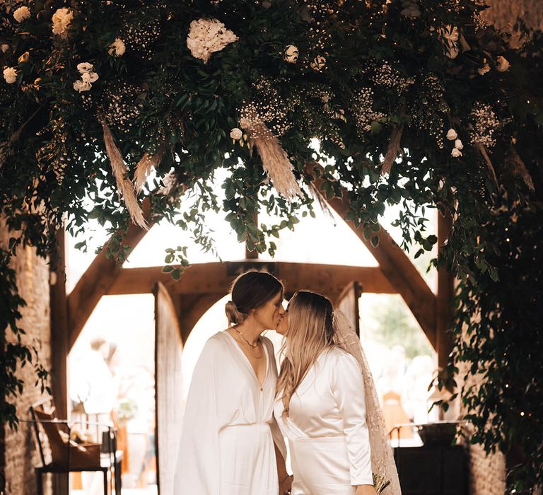 Brides kiss underneath floral installation on their wedding day