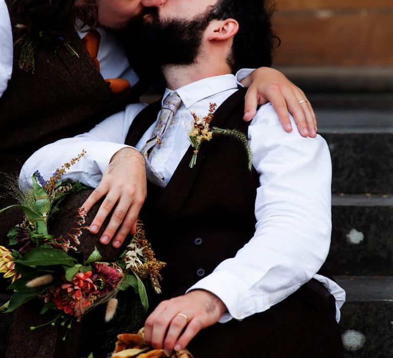 Grooms kiss on steps whilst holding their flower crowns in their laps