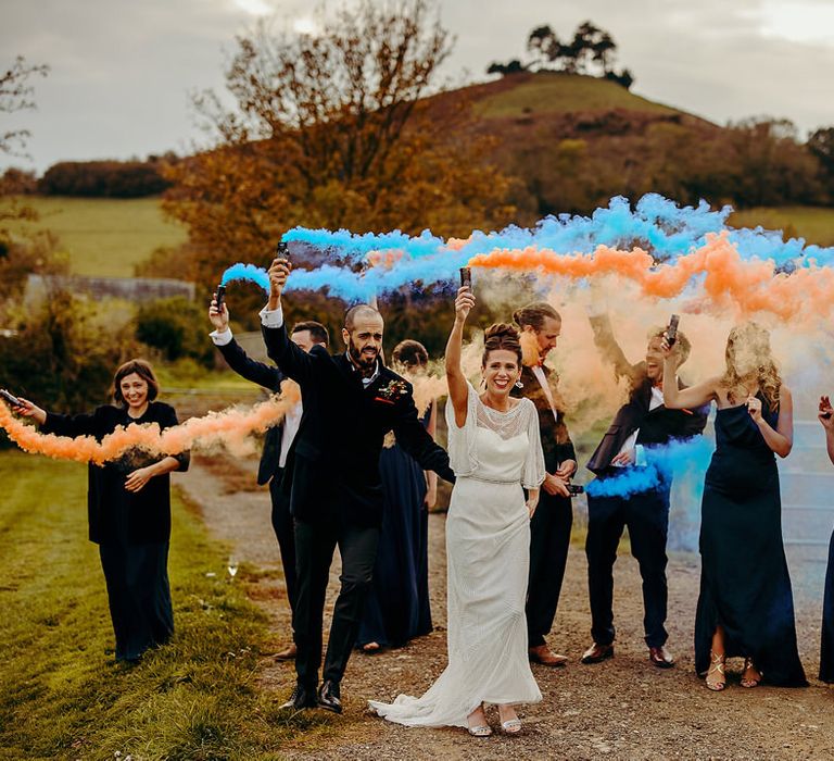 Wedding party smoke bomb photo with bridesmaids and groomsmen in navy and the bride in an embellished dress holding blue and orange grenades in the air 