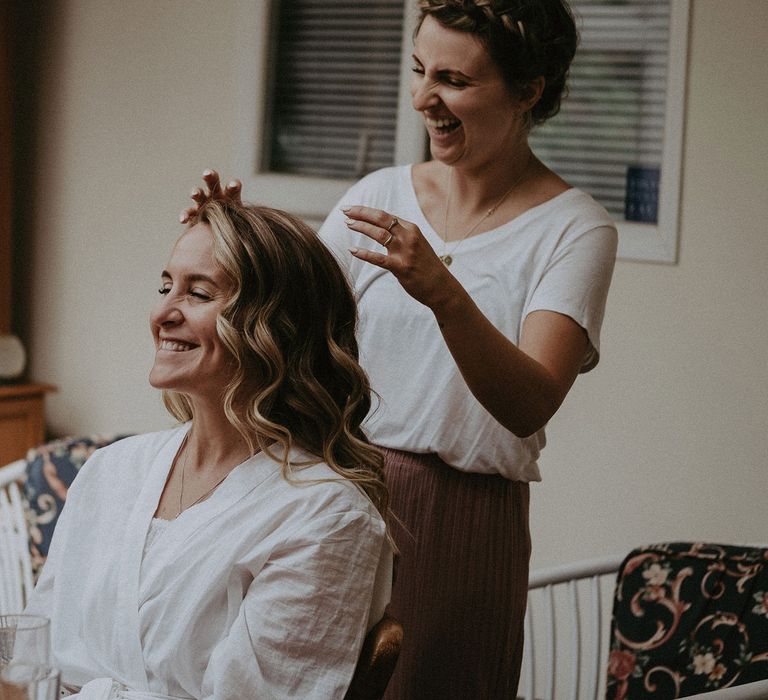 Bride with curled hair smiles as hairdresser arranges fringe sat in kitchen before Isle of Wight wedding with macrame wedding decor