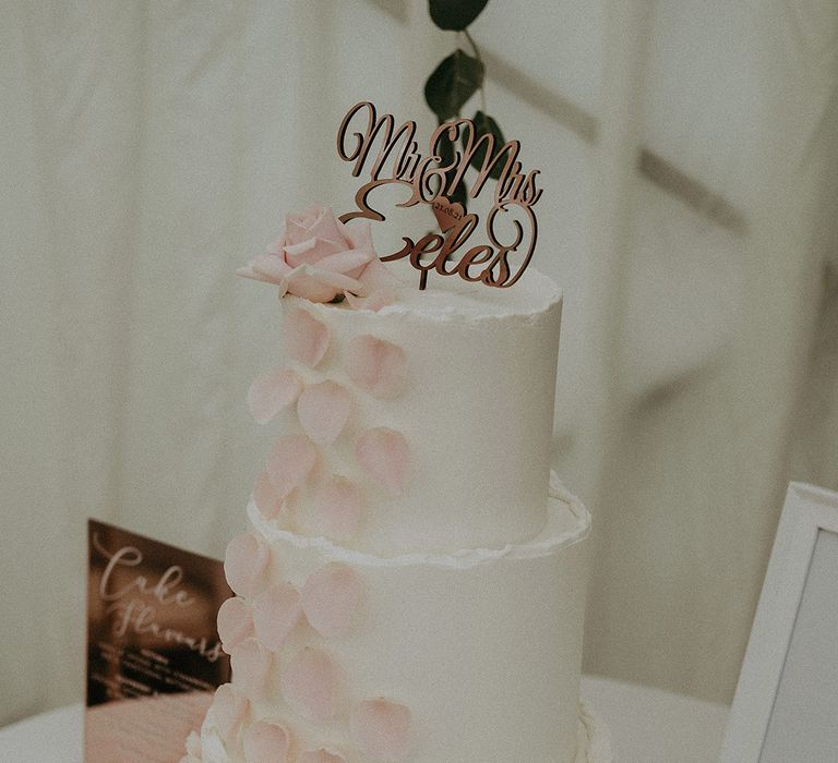 White three-tier wedding cake with pink petals cascading down the front and a personalised laser cut cake topper 