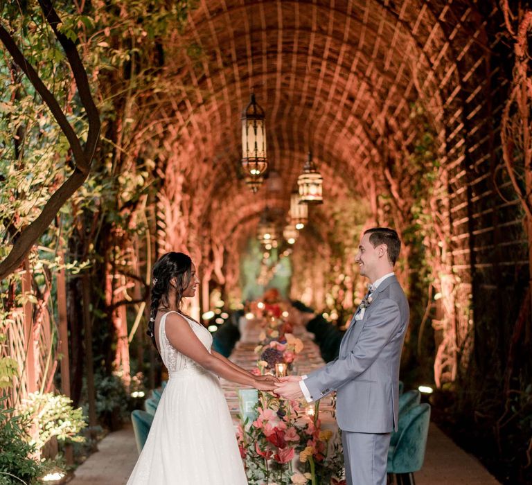 Bride & groom stand and hold hands whilst facing one another in front of reception table outdoors underneath floral pergola 