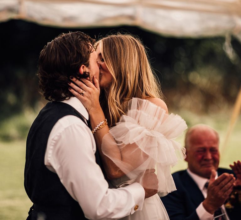 Groom in white shirt and blue waistcoat kisses bride in white Halfpenny London Mayfair dress during garden marquee wedding reception in Cornwall