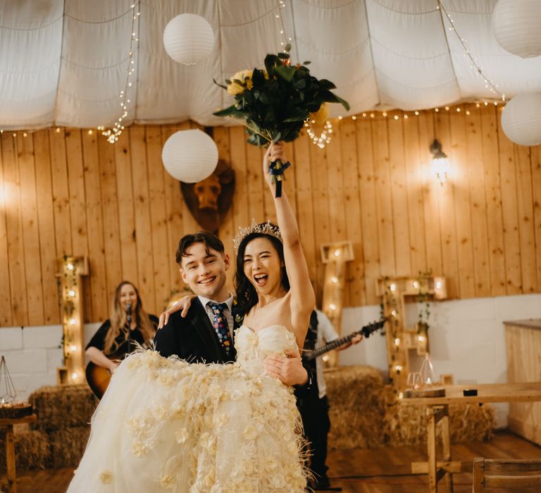 Groom in dark suit and floral tie carries bride in strapless feathered wedding dress and headpiece as she holds up bouquet during rustic wedding breakfast with fairy lights