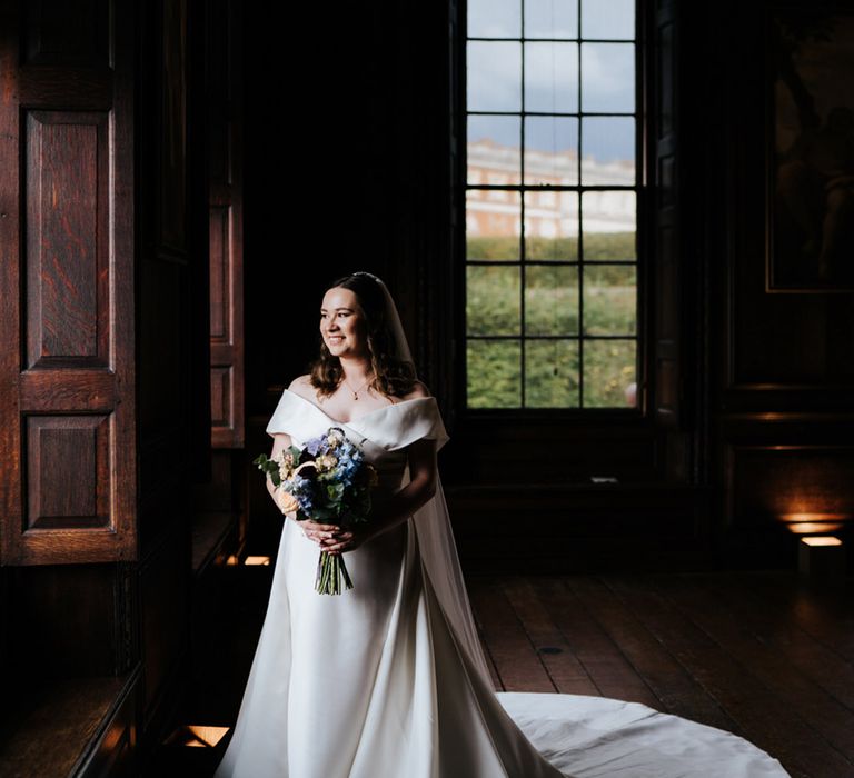 Bride stands in dark room in front of bright window as her long train spreads out around her