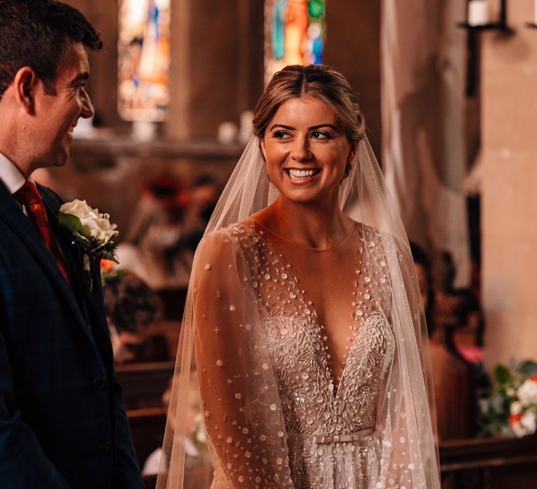 Bride in plunging v neck Julie Vino wedding dress and veil smiles at groom in checked blue suit and white buttonhole curing church wedding ceremony