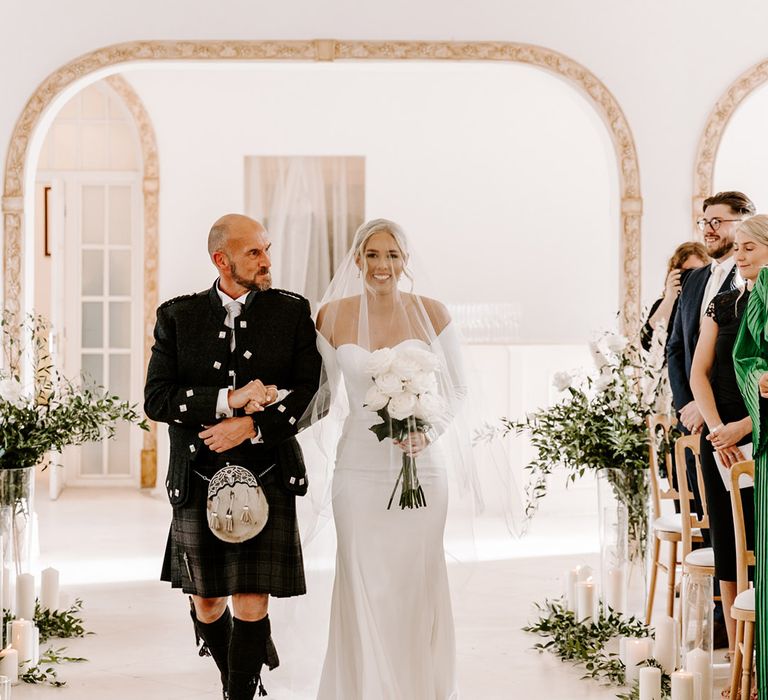 Bride walking down the aisle in a strapless wedding dress with long sleeves with her father in a tartan kilt and sporran 