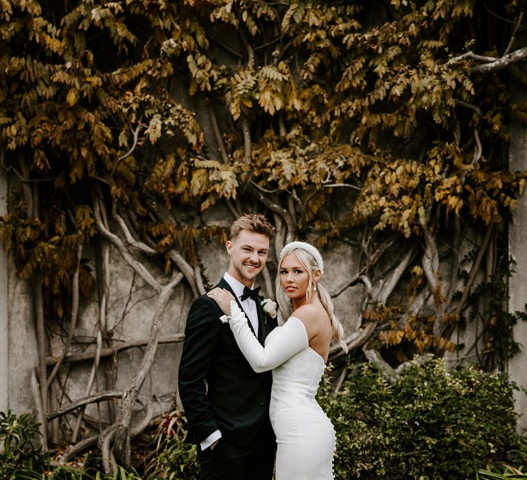 Stylish bride and groom in a dinner suit and minimalist fishtail wedding dress with buttons down the back and pearl headband 