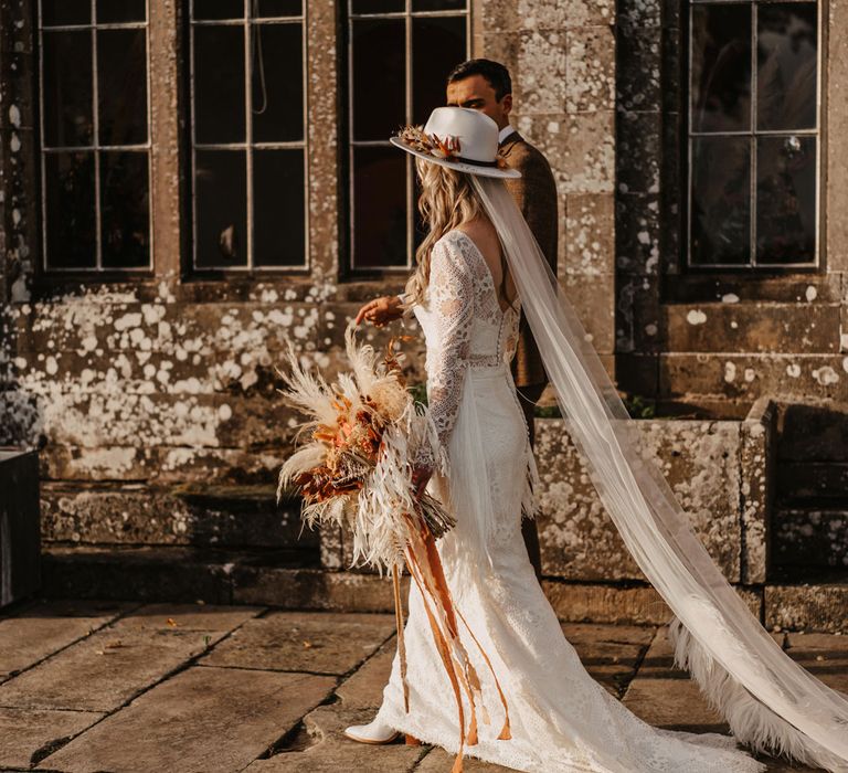 Bride in boho lace two piece wedding dress, bridal hat and fur trimmed veil by Rebecca Anne Designs holding white and orange boho wedding bouquet walks outside with groom in brown tweed suit 