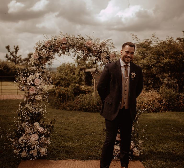 Groom in a three-piece suit waiting at the altar with a pink rose and white gypsophila flower arch 