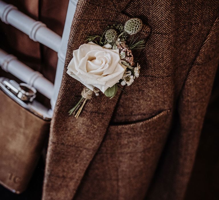 Brown tweed suit jacket hangs on chair with brown leather hipflask and white rose buttonhole