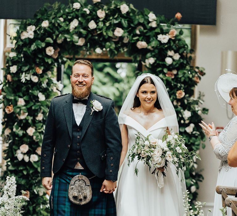 Bride in an off the shoulder Suzanne Neville wedding dress and groom in a tartan kilt standing in front of a floral arch