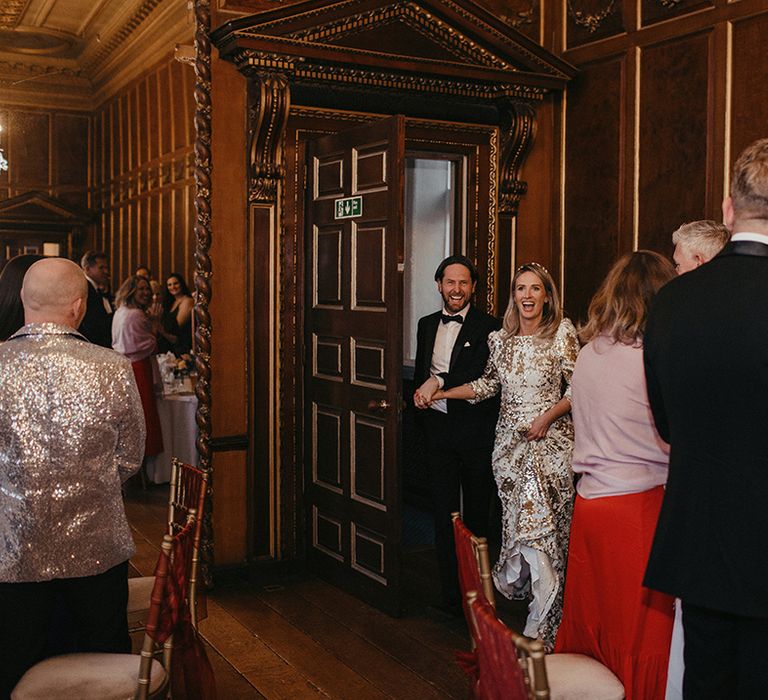 Bride walks into reception room with her groom whilst wearing sequinned Abigail Of Gardenia gown