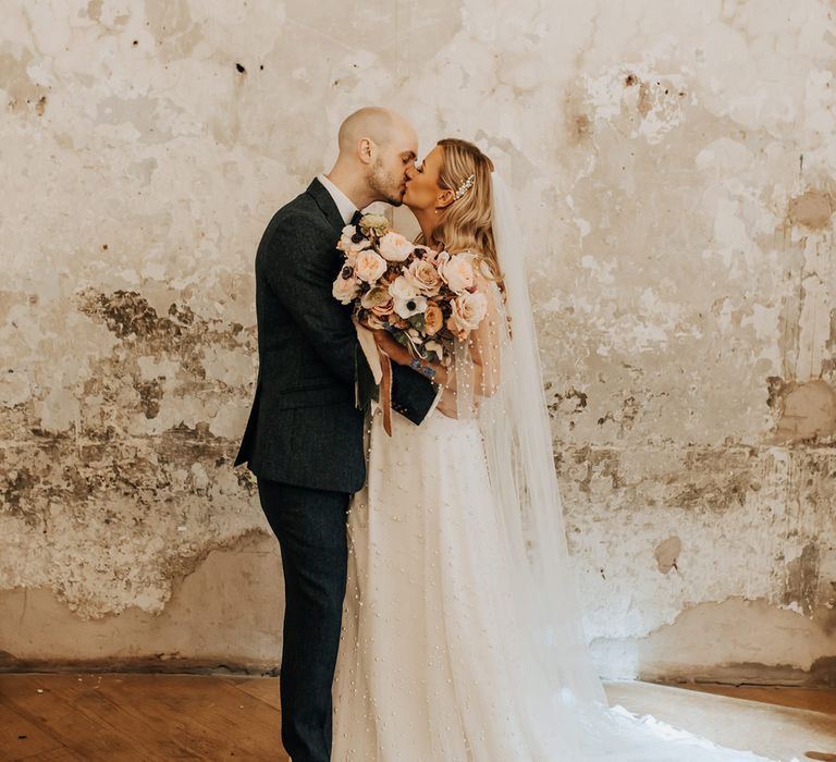 Bride in white wedding dress with pearl mesh overlay holding mixed rose and anemone bridal bouquet kisses groom at industrial wedding venue