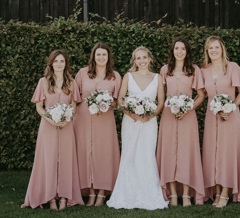 Bride in lace wedding dress with bridesmaids in muted pink button up dresses and high low skirt holding pink and white bouquets