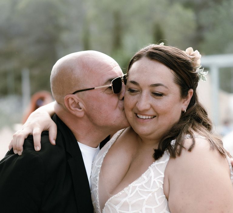 Bride with half up half down hair holding champagne flute gets a kiss on the cheek from a wedding guest 