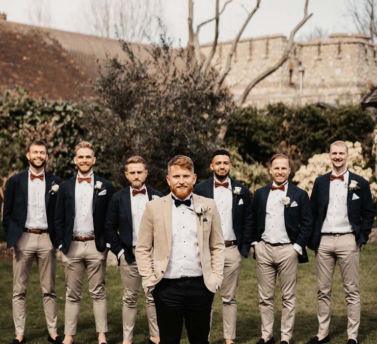 Groom in beige suit jacket and black bow tie and trousers and groomsman in light coloured trousers and orange bow ties