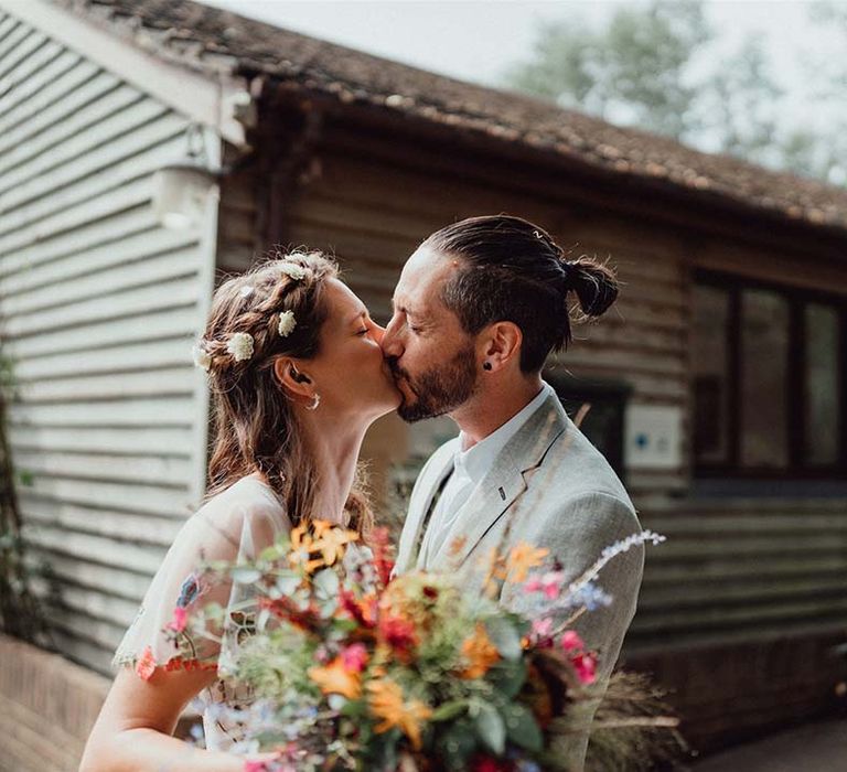 Bride and groom share a kiss outside their wedding venue with bride holding colourful wedding bouquet