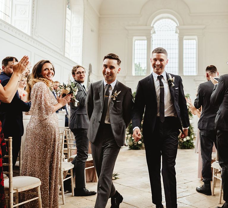 Grooms in grey and black suit with tie and gold tie clips walk back down the aisle as a married couple