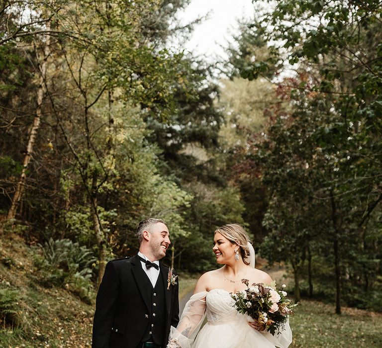 Bride and groom walk a path together holding hands with bride in custom wedding dress and groom in black tie and traditional kilt 