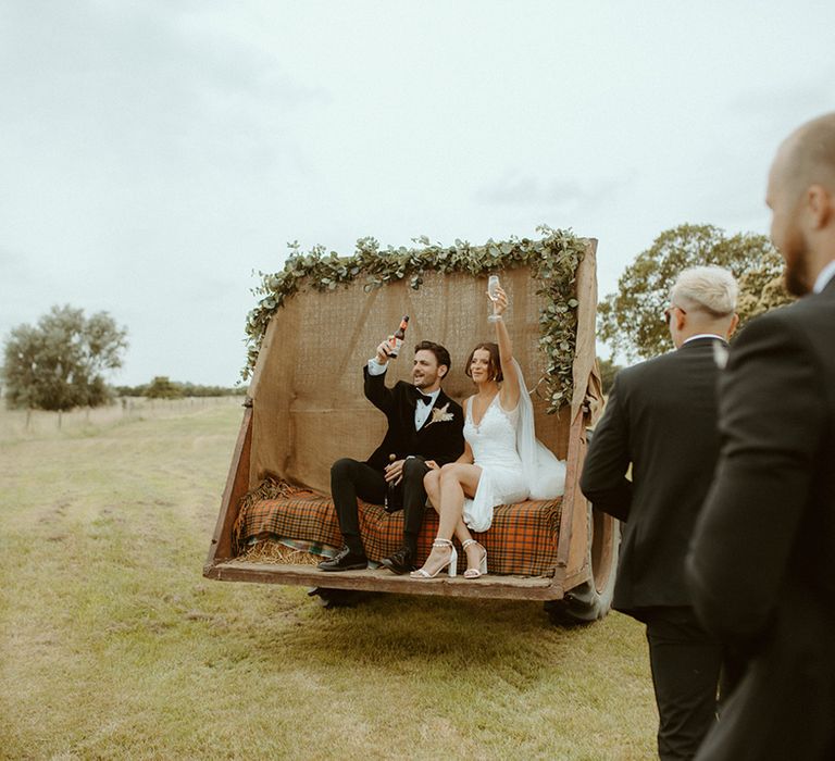 Bride and groom raise a toast to the bridesmaids and groomsmen as they sit on the back of a tractor 