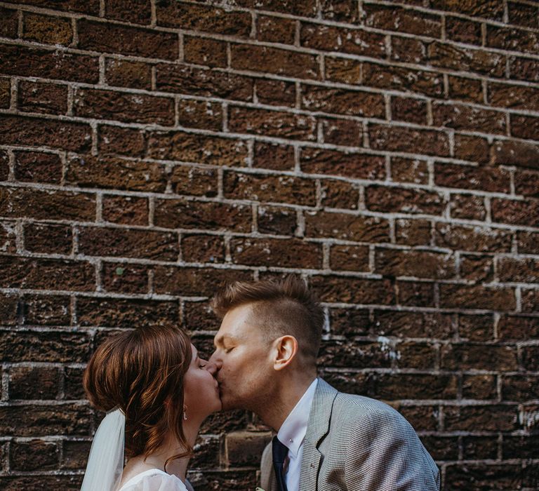 Bride and groom share a kiss in front of brick wall as bride holds spring wedding bouquet with giant poppy