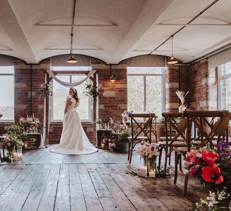 Bride in Jesus Peiro wedding dress with raised sleeves and white headband stands at the altar surrounded by bright spring flowers and wildflowers on crates and lit white pillar candles 