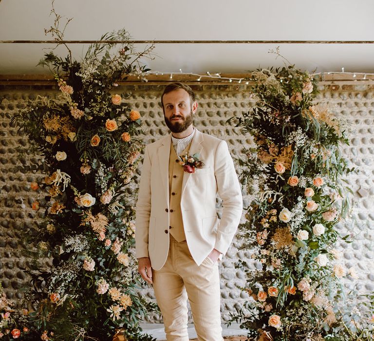 Groom in a three-piece beige suit standing in between column wedding flower arrangements made with blush pink and peach flowers and foliage 