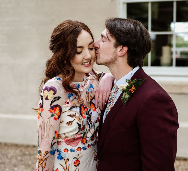 Groom kisses the bride on the cheek as she leans against him wearing bright flower wedding dress 