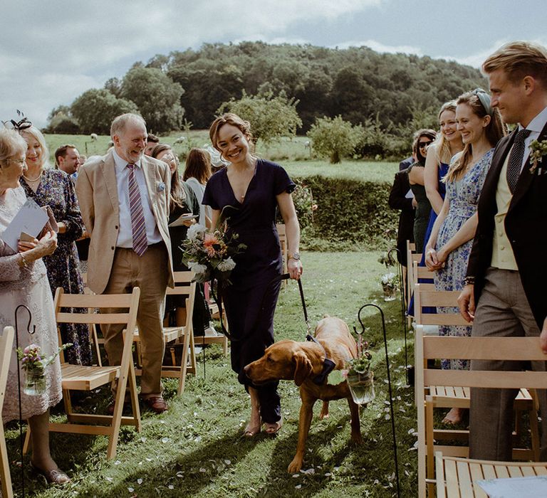 Bridesmaid in navy blue dress walks pet dog down the aisle 