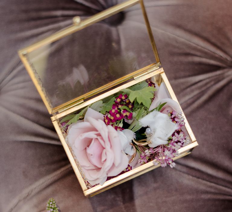 Wedding rings sit in gold and glass box surrounded by florals 