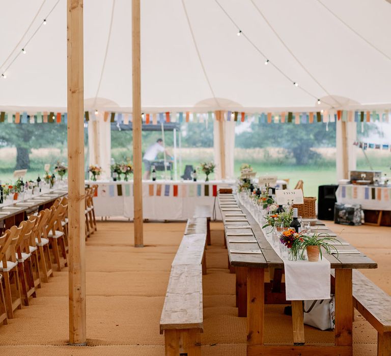 Colourful bunting across head table in marquee wedding complete with wooden banquet tables and festoon lighting 