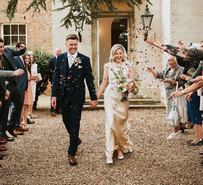 Bride wearing Kate Beaumont wedding gown in gold silk & groom in three piece suit walk through dried petal confetti outdoors 