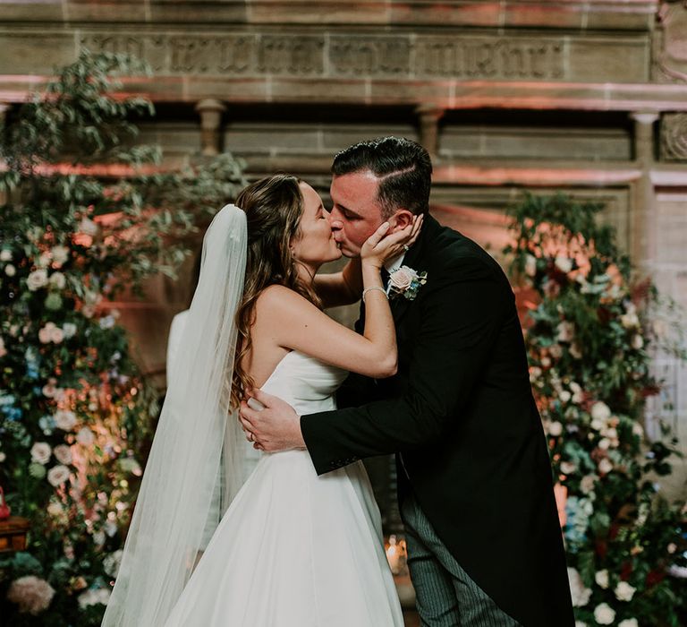 Bride wears Suzanne Neville princess wedding dress with sweetheart neckline whilst kissing her groom wearing morning suit and floral buttonhole