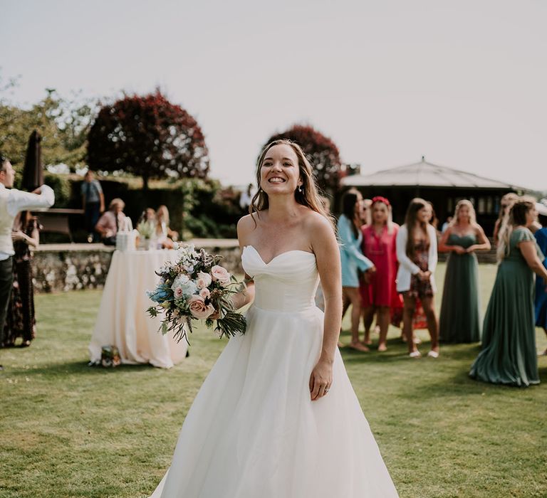 Bride wearing Suzanne Neville princess wedding dress holds pastel floral bouquet and walks through gardens at Cowdray House