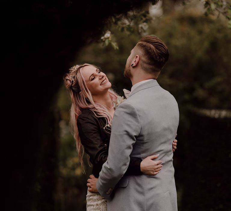 Bride wearing black denim jacket and wedding dress hugs her groom outdoors at Oxleaze Barn