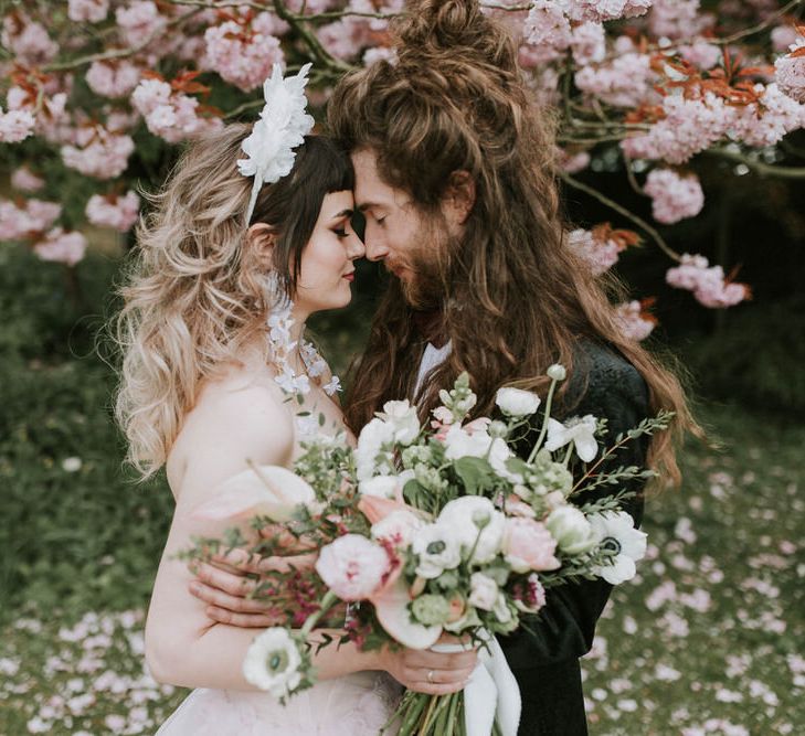 Couples' portrait under a blossom tree with bride in a white 3D flower headband and pink wedding dress and groom a black suit. 