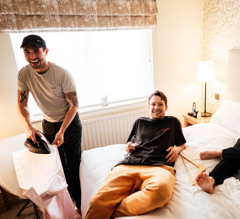 Groom lays on bed on the wedding morning as friends get ready before ceremony at Bradwell Hall 