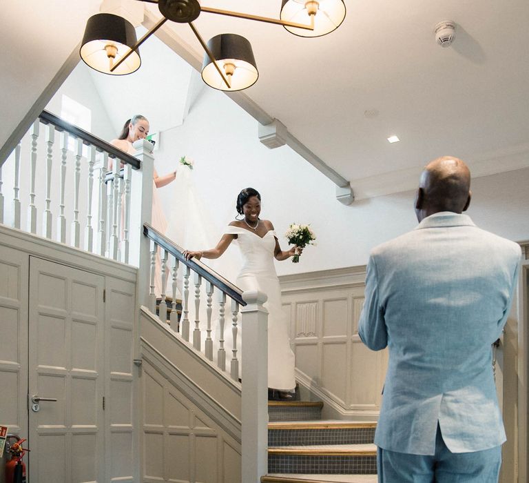 Bride walks down staircase carrying floral bouquet as father wearing pale blue suit sees her for the first time