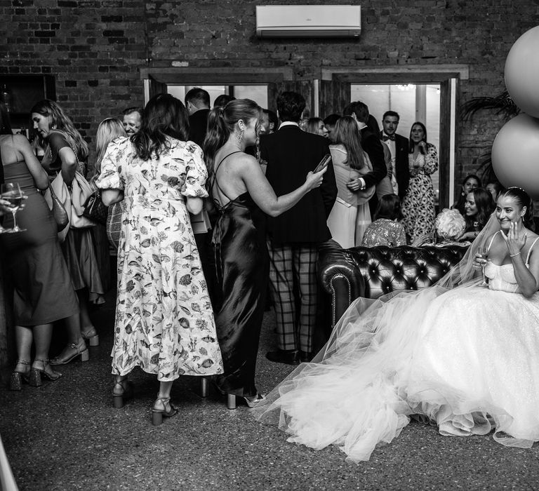 Bride wearing sparkly wedding dress with princess skirt sits on leather sofa at the 100 Barrington as bridesmaid in black bridesmaid dress takes picture of her 