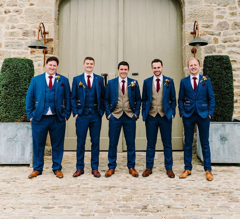 Groom stands with his groomsmen who wear three piece suits with blue suit jackets maroon ties and tartan waistcoats 