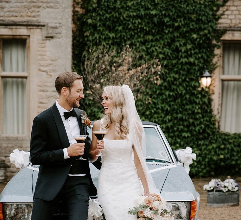 Bride and groom lean against their vintage blue car with glasses of espresso martinis 