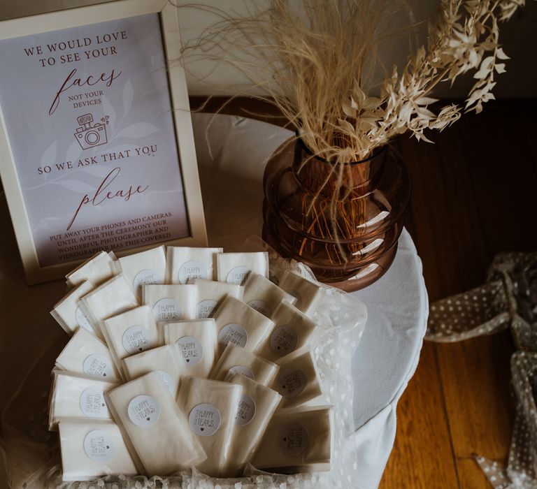 Unplugged wedding ceremony in a white frame with small white packets of tissues with "For your happy tears" written on them by a vase with a dried flower arrangement 