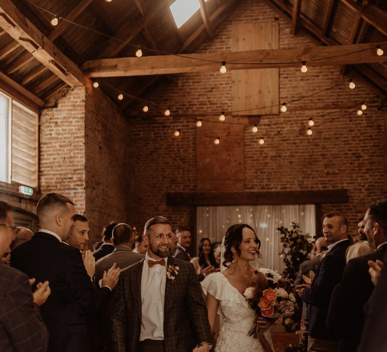 Festoon wedding lighting decorate the rustic barn wedding venue as the bride and groom walk back down the aisle as a married couple